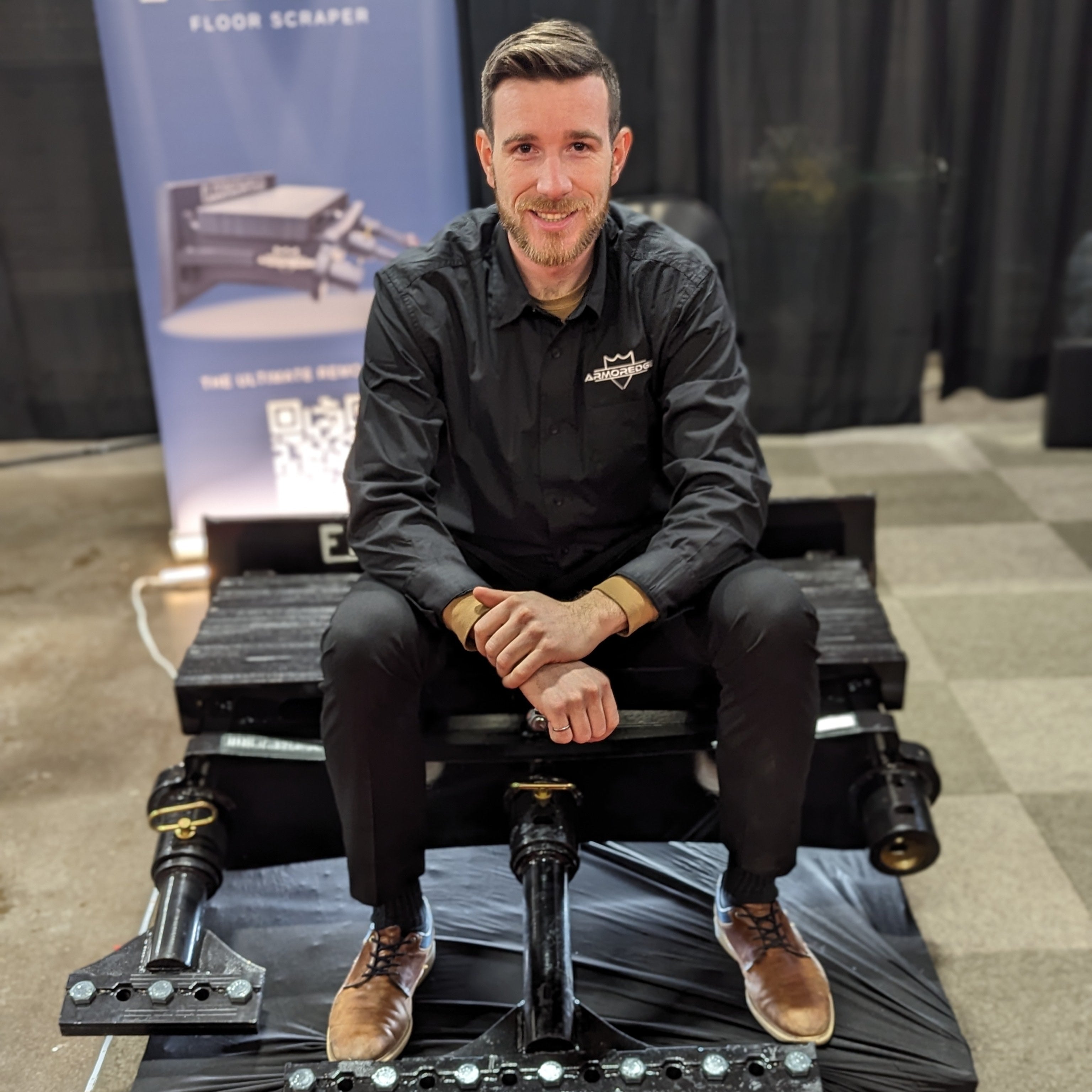 Man sitting on a large floor scraper at an event with a banner in the background.